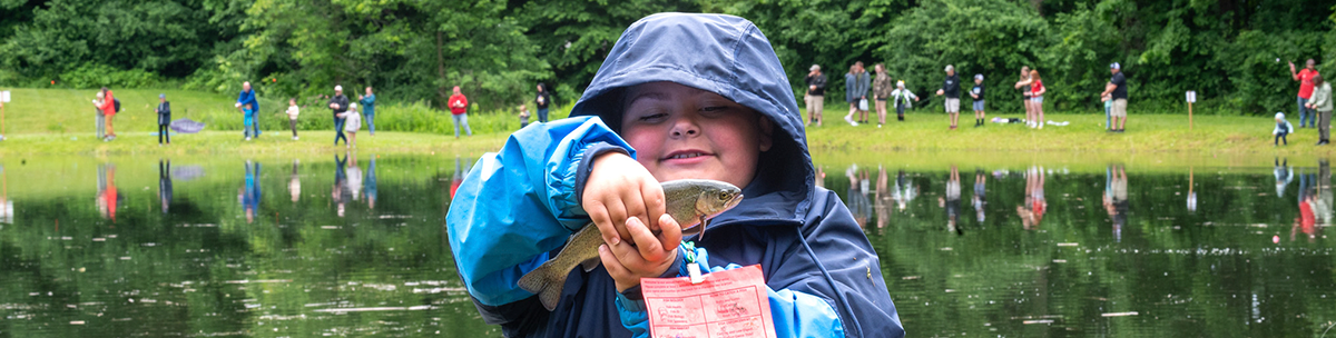 rosy cheeked child in raincoat shows off the rainbow trout he caught with more folks fishing in the distance at the pond behind him