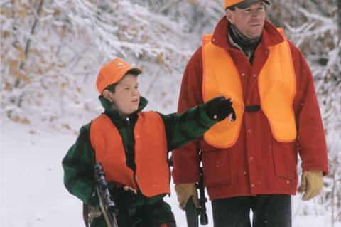 Boy pointing off camera to his hunting mentor in snowy woods, both wearing blaze orange vests and hat.